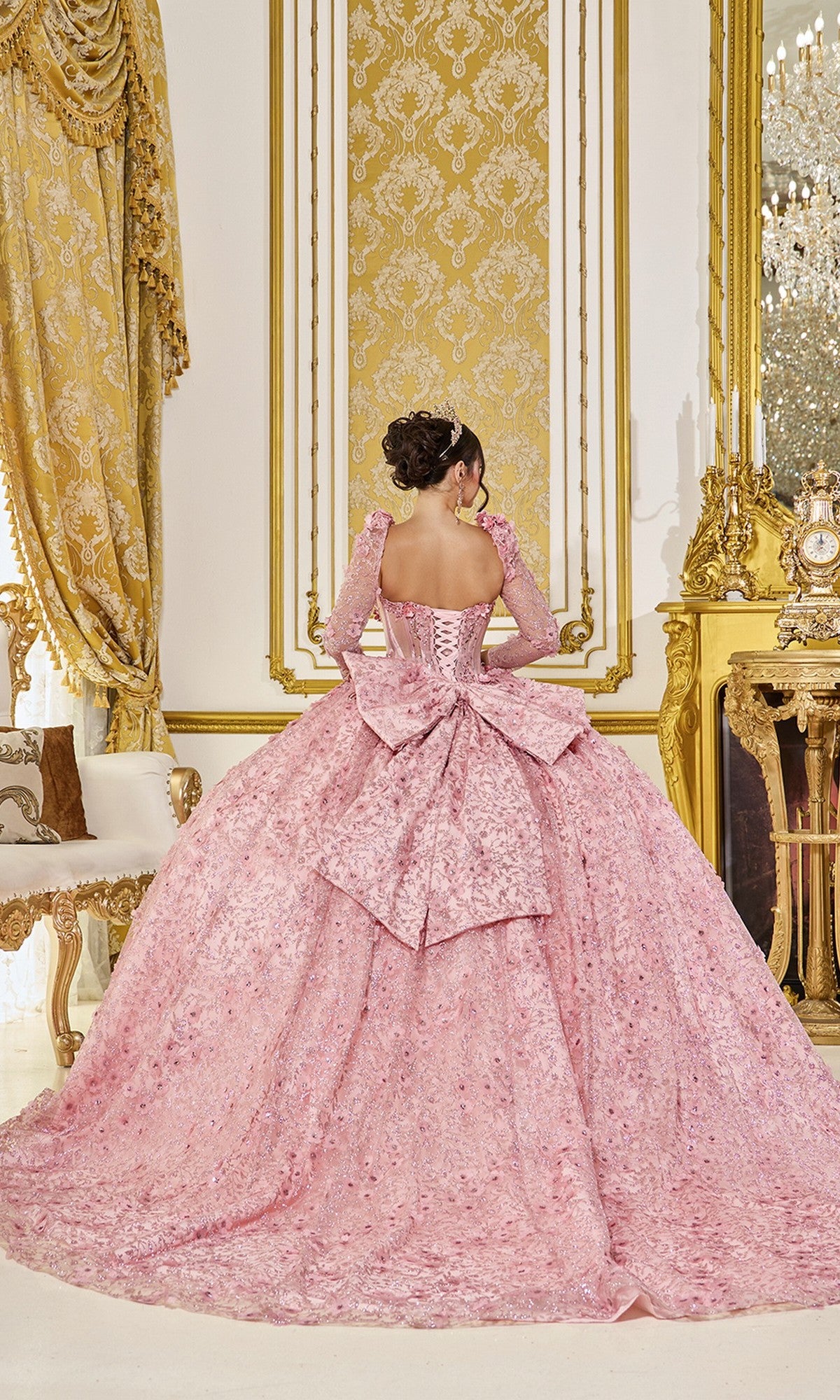 A woman wearing the Quinceanera Dress 5077 By Dancing Queen, featuring a blush pink corset bodice and large bow, stands facing away in an opulent room with gold accents and elaborate decor.